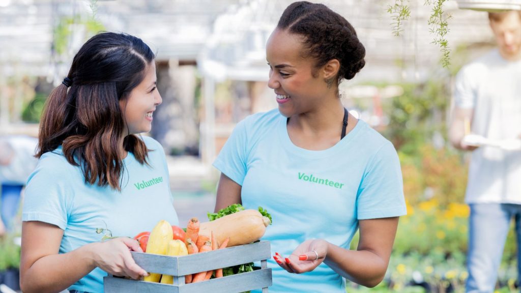 Two women wearing 'Volunteer' t-shirts holding a box of vegetables in a garden.