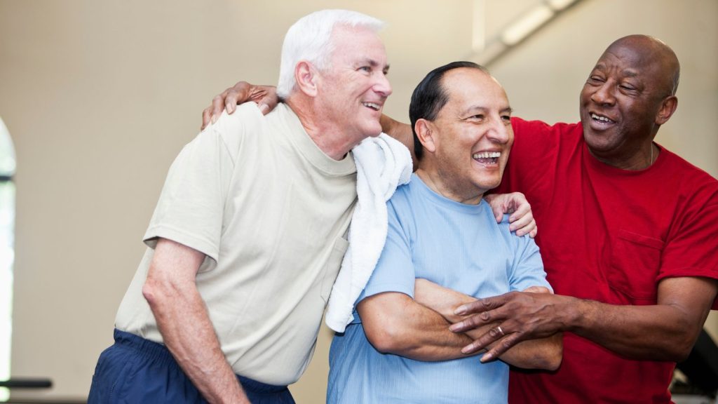 Three men aged around 60 laughing and smiling with their arms round each other.