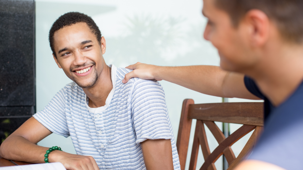 A young man smiling. His friend has a supportive hand on his shoulder.