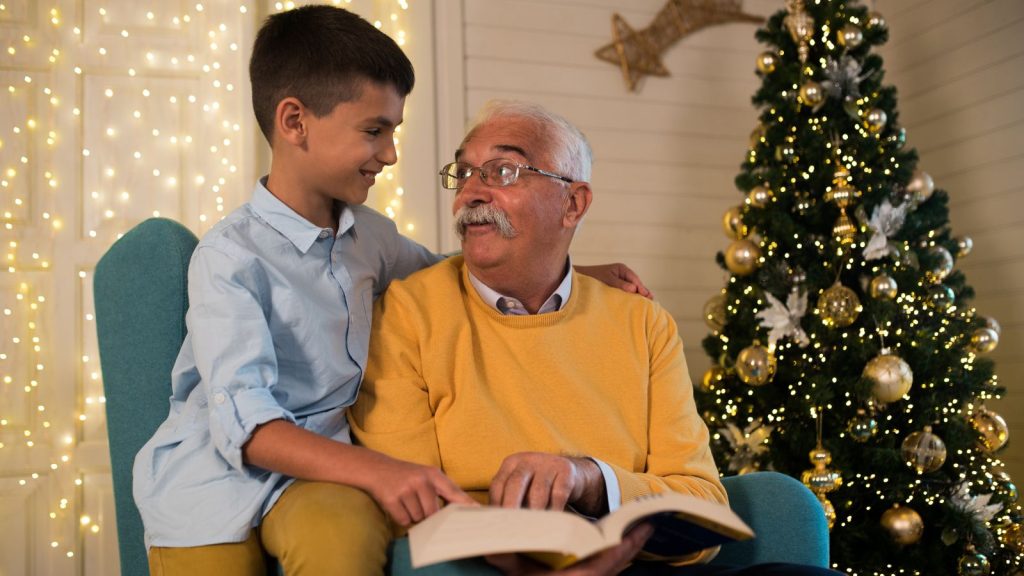 A young boy reading a book with his grandfather, with a Christmas tree in the background.