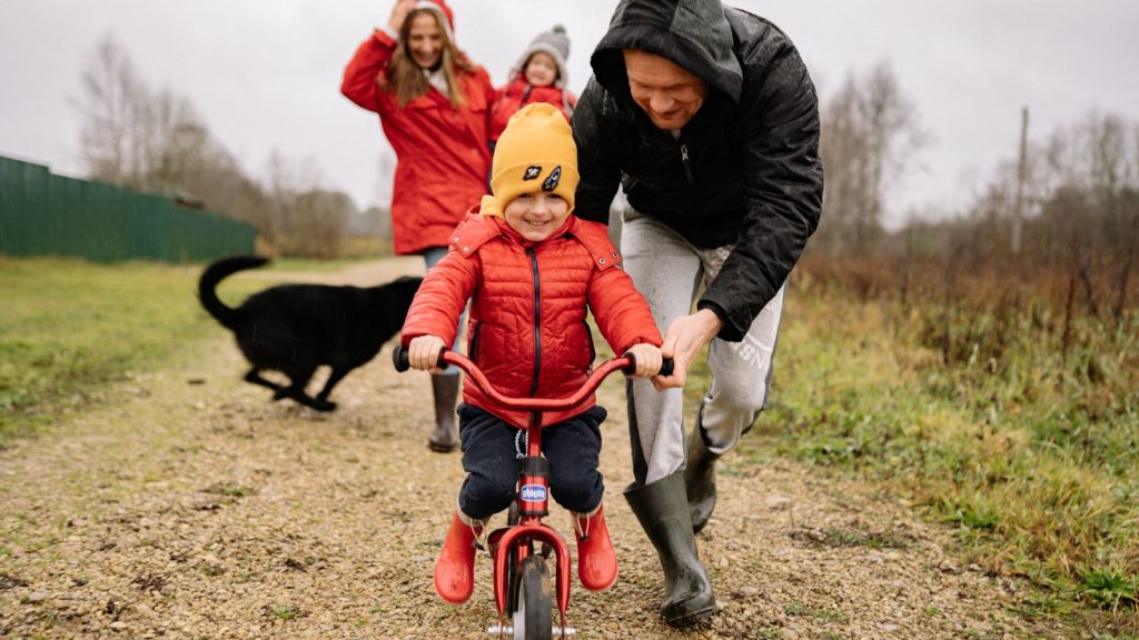 A father helping his son ride a bike while the rest of the family walks behind.