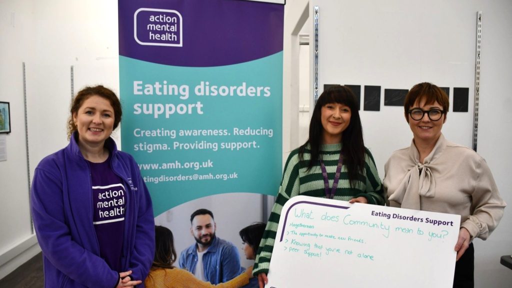Deborah, Lorraine and Shauna stand beside an Action Mental Health banner, holding a board which reads 'What does community mean to you?
