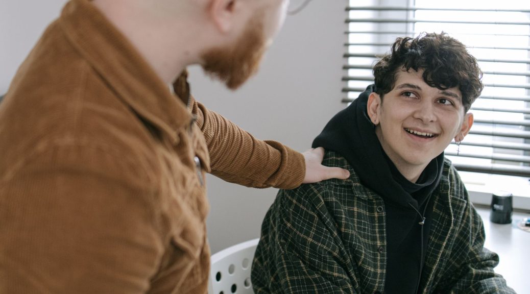 A man stands with his hand on another young man's shoulder in a show of support.