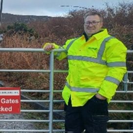 Sam stands by a gate wearing a hi-vis jacket, in his training placement role.