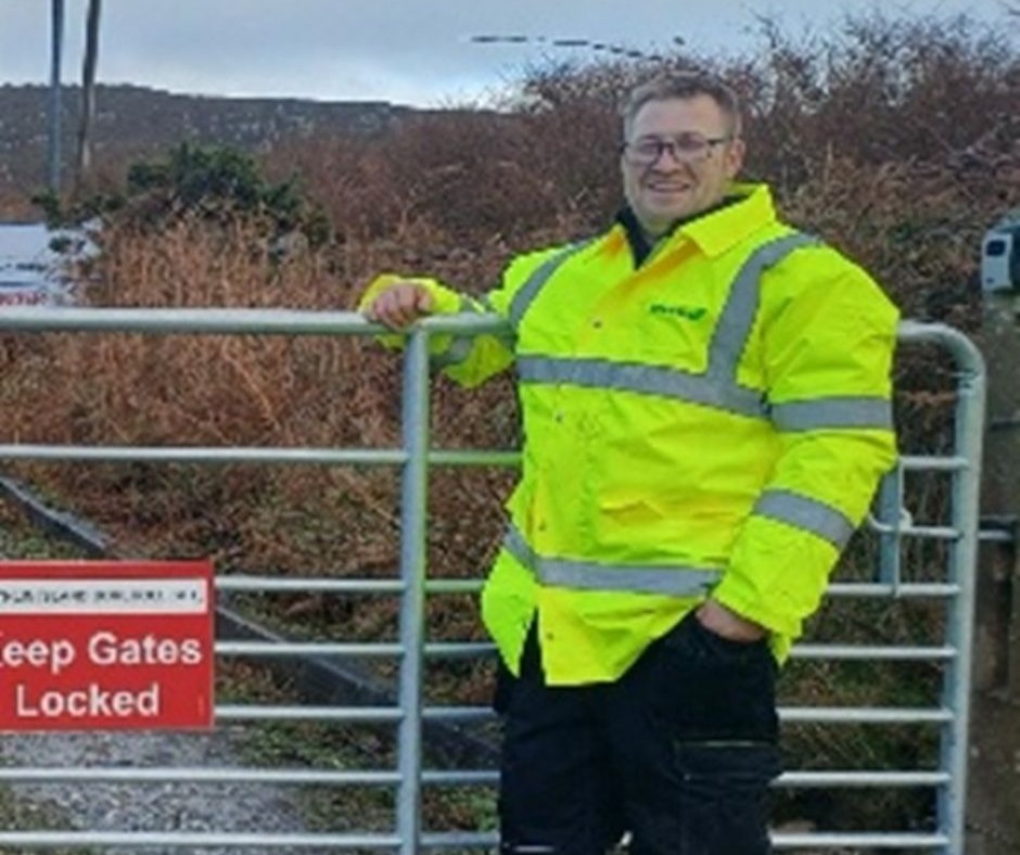 Sam stands by a gate wearing a hi-vis jacket, in his training placement role.
