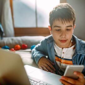 A teenage boy scrolling on his phone while sitting by a laptop,