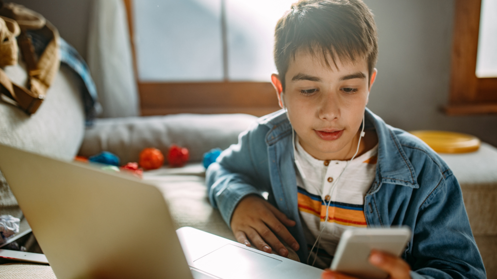 A young teenage boy scrolling on a mobile phone while sat in front of a laptop.